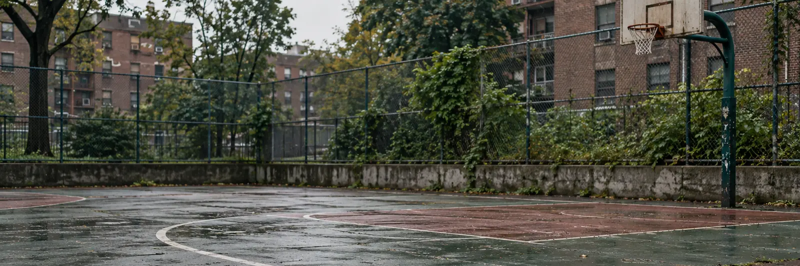 An empty outdoor basketball court after rain, cracked and wet, the hoop standing alone by a chain link fence